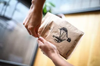 Ruka otevírá hnědý papírový sáček kávy se černou ilustrací ptáka