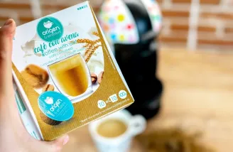 Ruka drží krabičku kapslí Origin Café con avena kávovar Dolce Gusto v pozadí šálek kávy