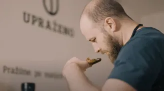 Muž s plnovousem v tmavém tričku skloněný nad nástrojem pro kávu v pražírně na stěně viditelný nápis