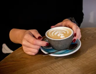 Ruce s červenými nehty drží šálek kávy s latte art na dřevěném stole