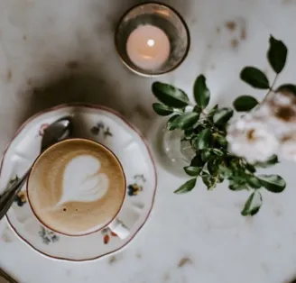 kávový šálek s latte artem ve tvaru srdce na podšálku s květinami a svíčkou