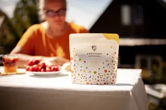 Pytlík kávy s barevným vzorem na stole venku v pozadí člověk v oranžovém tričku a talíř s ovocem