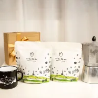 Dvě bílé sáčky UPRAŽENO s zeleným motivem krajiny na spodní části stojí na stole vedle moka konvičky a hrníčku