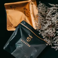 dvě balené zrnkové kávy modrý sáček s nápisem ROASTED zlaté balení vzadu suché květy