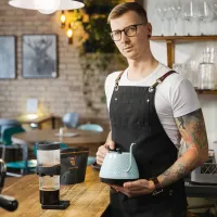 Barista v bílé tričko a černé zástěře drží světlomodrou konvičku na kávu u dřevěného pultu kavárny