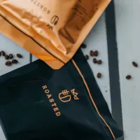 zrnková káva dvě balení tmavě modrý a béžový obal s logem Roasted kolem rozptýlených zrn kávy