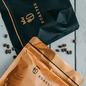 balení kávy černé a měděné barvy s logem ROASTED a rozloženými zrny kávy