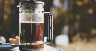 Skleněný French press s černým rámem stojí na venkovním stole s rozostřeným přírodním pozadím