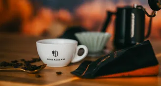 Bílý šálek s logem Uprazeno na dřevěném stole vedle balíčku zrnkové kávy a kávového příslušenství v pozadí