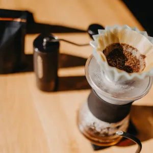 Papírový filtr s namletou kávou na kovovém dripperu nad skleněnou konvičkou na dřevěném stole v pozadí