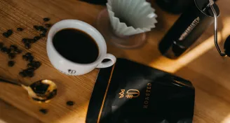 Šálek černé kávy na dřevěném stole vedle balíčku kávy s nápisem ROASTED a zrna kávy kolem