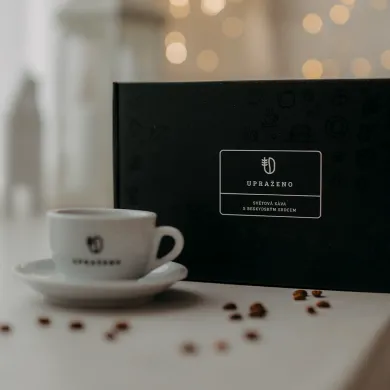 černé dárkové balení s bílým logem UPRAŽENO šálek s podšálkem v popředí a rozptýlená zrna kávy