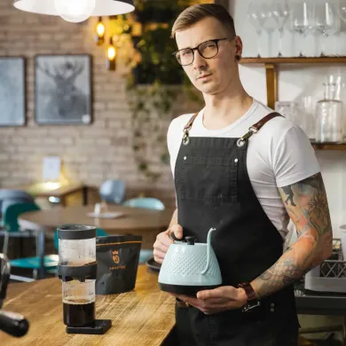 barista-v-bile-tricko-a-cerne-zastere-drzi-svetlomodrou-konvicku-na-kavu-u-dreveneho-pultu-kavarny